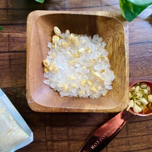 May include: A wooden square bowl filled with white bath salts and dried flower petals. A copper measuring spoon with the word "CROFTON" is next to the bowl. A small bag of bath salts is in the background. Green leaves are in the background.