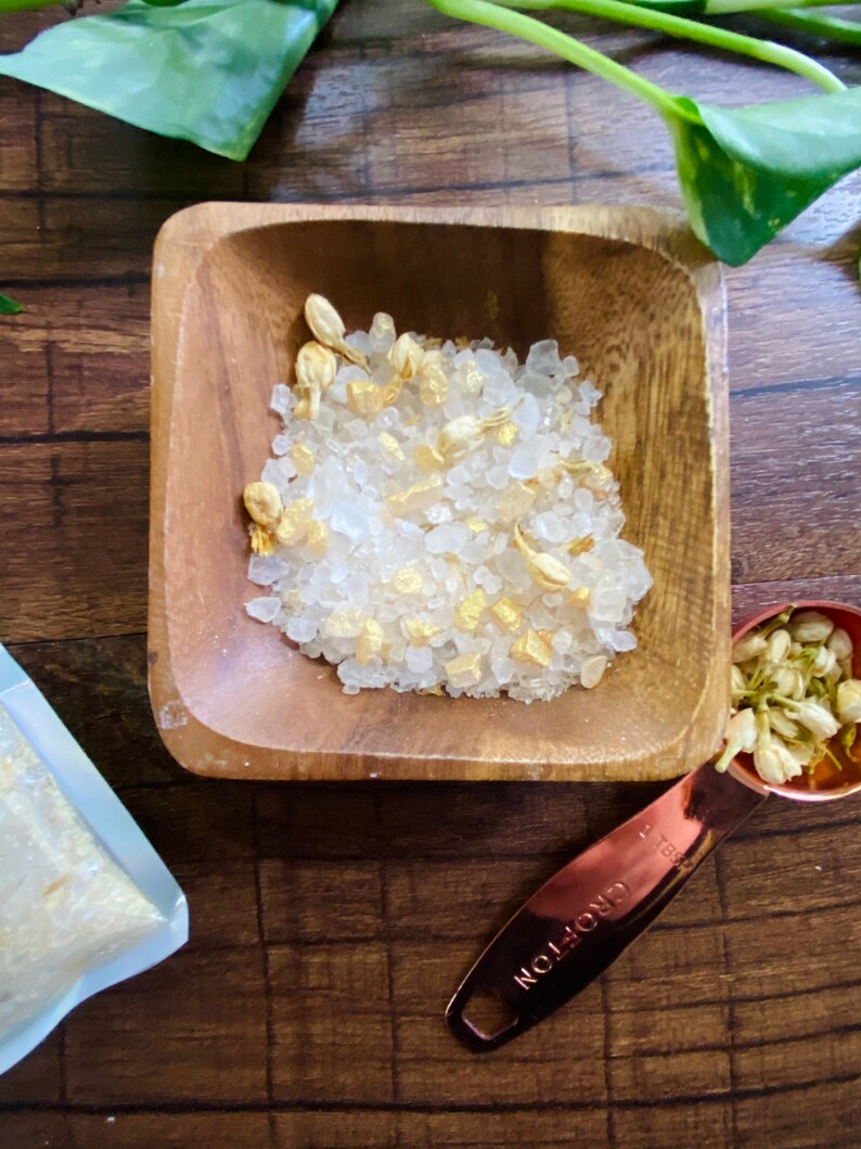 May include: A wooden square bowl filled with white bath salts and dried flower petals. A copper measuring spoon with the word "CROFTON" is next to the bowl. A small bag of bath salts is in the background. Green leaves are in the background.