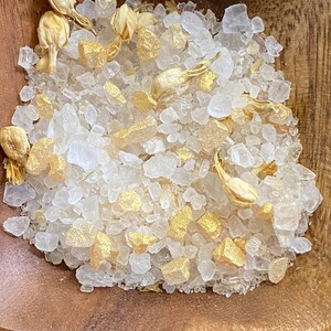May include: A wooden bowl containing bath salts. The salts are composed of large, clear crystals, gold-coloured chunks, and dried jasmine buds. The image is a close-up, emphasising the textures and colours of the ingredients.