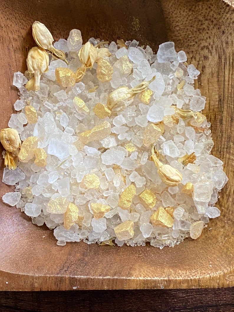 May include: A wooden bowl containing bath salts. The salts are composed of large, clear crystals, gold-coloured chunks, and dried jasmine buds. The image is a close-up, emphasising the textures and colours of the ingredients.