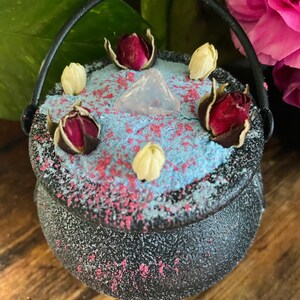 Könnte beinhalten: Ein kleiner schwarzer Kessel mit Henkel, gefüllt mit blauen und rosa Badesalzen, getrockneten Rosenknospen und einem klaren Quarzkristall.