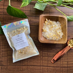 May include: A clear, holographic pouch and a small wooden bowl filled with bath salts and dried jasmine flowers. A copper measuring spoon holds more jasmine. The pouch reads "The Alchemist Detox Bath" with ingredients listed. The products are arranged on a dark wood surface.