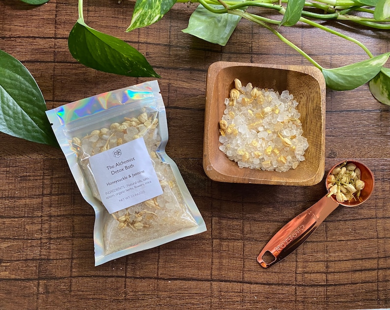 May include: A clear, holographic pouch and a small wooden bowl filled with bath salts and dried jasmine flowers. A copper measuring spoon holds more jasmine. The pouch reads "The Alchemist Detox Bath" with ingredients listed. The products are arranged on a dark wood surface.