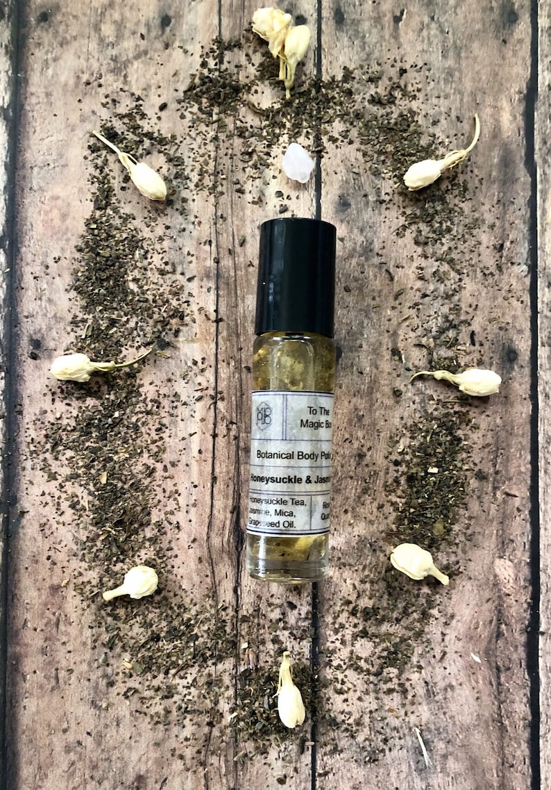 May include: A small glass bottle with a black rollerball top and a white label. The label reads "To The Magic Ba Botanical Body Pol Honeysuckle & Jasm Honeysuckle Tea, Jame, Mica, Grapeseed Oil."