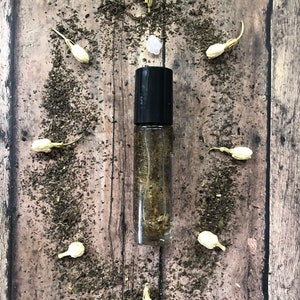 May include: A clear glass roller bottle with a black cap filled with a golden liquid. The bottle is surrounded by small white flowers and green tea leaves on a wooden surface.