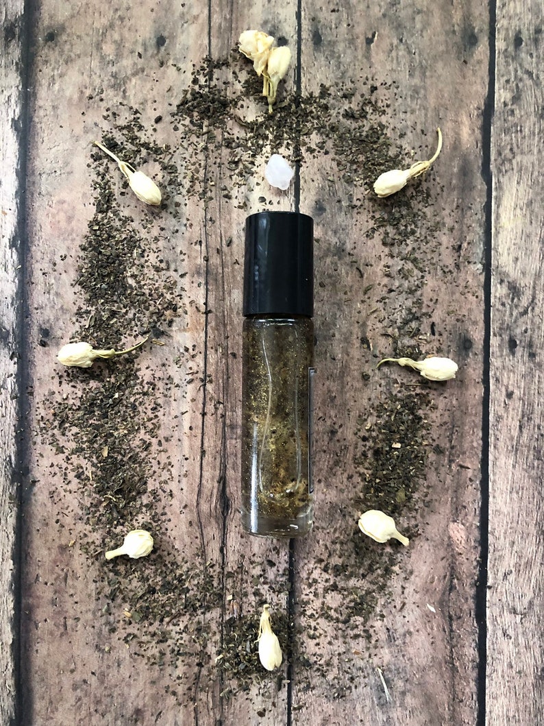 May include: A clear glass roller bottle with a black cap filled with a golden liquid. The bottle is surrounded by small white flowers and green tea leaves on a wooden surface.