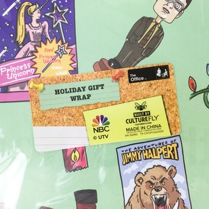 May include: A corkboard with a white note that says "HOLIDAY GIFT WRAP" and a smaller note that says "BUILT BY CULTUREFLY" with the company website and "MADE IN CHINA". There is a small NBC logo and a sticker that says "The Office."