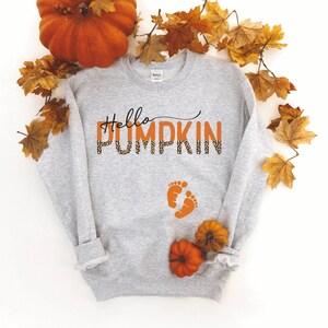 K&ouml;nnte beinhalten: Grauer Pullover mit dem Text "Hello Pumpkin" in Orange mit Leopardenmuster. Der Text ist in einer kursivschrift. Unter dem Text befinden sich zwei orangefarbene Fu&szlig;abdr&uuml;cke. Der Pullover ist von Herbstbl&auml;ttern und einem K&uuml;rbis umgeben.