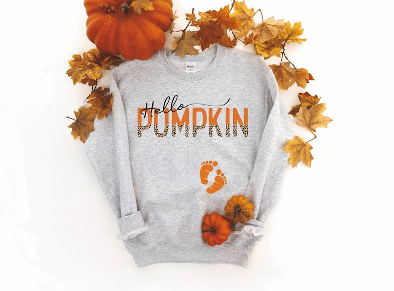 K&ouml;nnte beinhalten: Grauer Pullover mit dem Text "Hello Pumpkin" in Orange mit Leopardenmuster. Der Text ist in einer kursivschrift. Unter dem Text befinden sich zwei orangefarbene Fu&szlig;abdr&uuml;cke. Der Pullover ist von Herbstbl&auml;ttern und einem K&uuml;rbis umgeben.