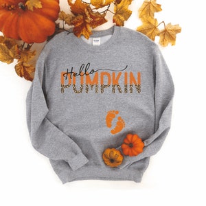K&ouml;nnte beinhalten: Grauer Pullover mit dem Text "Hello Pumpkin" in Orange mit Leopardenmuster. Der Text ist fett gedruckt. Unter dem Text befinden sich zwei orangefarbene Babyf&uuml;&szlig;e.