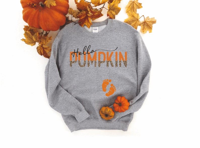 K&ouml;nnte beinhalten: Grauer Pullover mit dem Text "Hello Pumpkin" in Orange mit Leopardenmuster. Der Text ist fett gedruckt. Unter dem Text befinden sich zwei orangefarbene Babyf&uuml;&szlig;e.