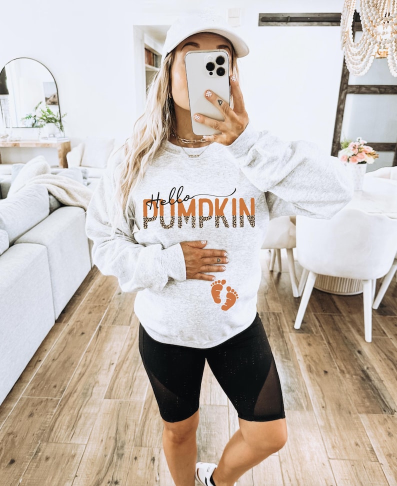 K&ouml;nnte beinhalten: Ein graues Sweatshirt mit dem Text "Hello Pumpkin" in orangefarbener und schwarzer Schrift. Das Sweatshirt hat au&szlig;erdem zwei orangefarbene Baby-Fu&szlig;abdr&uuml;cke. Die Person, die das Sweatshirt tr&auml;gt, ist schwanger.