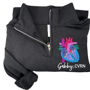May include: Black quarter-zip sweatshirt with a colorful anatomical heart graphic and the text "Gabby, CVRN".
