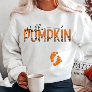 K&ouml;nnte beinhalten: Wei&szlig;er Pullover mit dem Text "Hello Pumpkin" in orangefarbenem und schwarzem Leopardenmuster mit zwei orangefarbenen Babyf&uuml;&szlig;chen darunter.