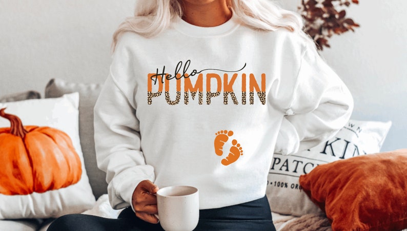 K&ouml;nnte beinhalten: Wei&szlig;er Pullover mit dem Text "Hello Pumpkin" in orangefarbenem und schwarzem Leopardenmuster mit zwei orangefarbenen Babyf&uuml;&szlig;chen darunter.