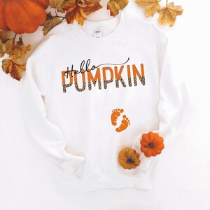 K&ouml;nnte beinhalten: Wei&szlig;er Pullover mit dem Text "Hello Pumpkin" in orangefarbenem und schwarzem Leopardenmuster. Der Text ist mit zwei kleinen orangenen Fu&szlig;abdr&uuml;cken darunter &uuml;berlagert.