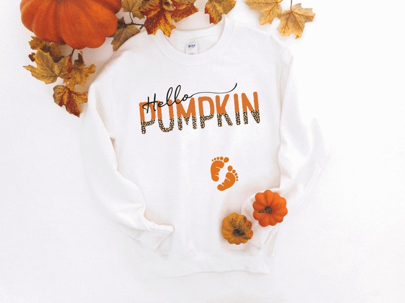 K&ouml;nnte beinhalten: Wei&szlig;er Pullover mit dem Text "Hello Pumpkin" in orangefarbenem und schwarzem Leopardenmuster. Der Text ist mit zwei kleinen orangenen Fu&szlig;abdr&uuml;cken darunter &uuml;berlagert.