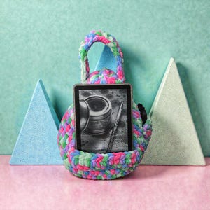 May include: A colorful, hand-crocheted tablet sleeve with a handle, holding a tablet displaying a black and white image of a pen and inkwell. The sleeve is made of chunky yarn in shades of pink, green, blue, and purple.