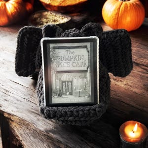 May include: A black crocheted book holder with a book titled "The Pumpkin Spice Cafe" by Laurie Gilmore. The book holder is sitting on a wooden table with a lit candle and pumpkins in the background.