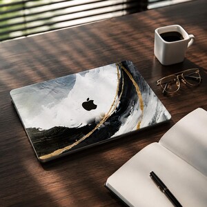 May include: A laptop skin featuring an abstract design with sweeping strokes of black, white, and gray paint, accented by a thin, textured gold line running diagonally across the surface, centered with a black Apple logo cutout.