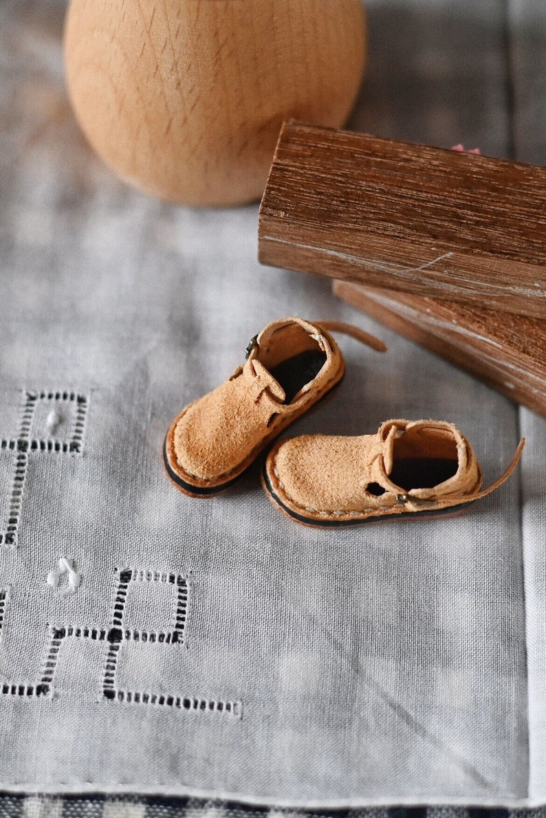 K&ouml;nnte beinhalten: Ein Paar Miniatur-Sandalen aus braunem Leder mit schwarzen N&auml;hten und Schnallen. Die Sandalen liegen auf einer wei&szlig;en Tischdecke aus Leinen mit einem dekorativen Muster.