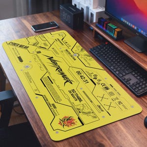 May include: A yellow rectangular desk mat with black graphic designs and text, including "CYBER WEAR" and "NIGHTRUNNER". The mat features a digital clock display and a stylized dragon graphic. The mat is on a wooden desk.