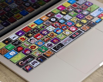 May include: A laptop keyboard skin featuring colorful, square stickers with various cartoon and video game character designs. The skin covers the keys with a variety of images, including a retro game controller. The text "scanace MACBOOK SKIN" is visible at the top.