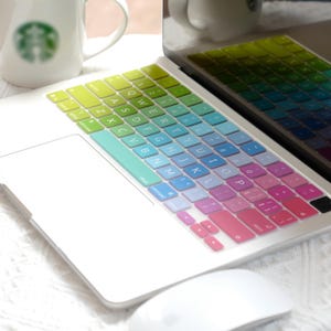 May include: A laptop computer featuring a vibrant, multi-colored keyboard cover with a gradient transition from lime green on the left side to bright pink on the right side. The QWERTY layout is clearly visible on the keys, which are set against a white laptop body.