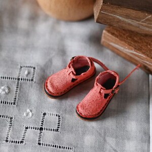 K&ouml;nnte beinhalten: Ein Paar Miniatur-Pinkschuhe aus Leder mit Schnallen. Die Schuhe befinden sich auf einer wei&szlig;en Stoffoberfl&auml;che mit einem zarten Stickmuster.