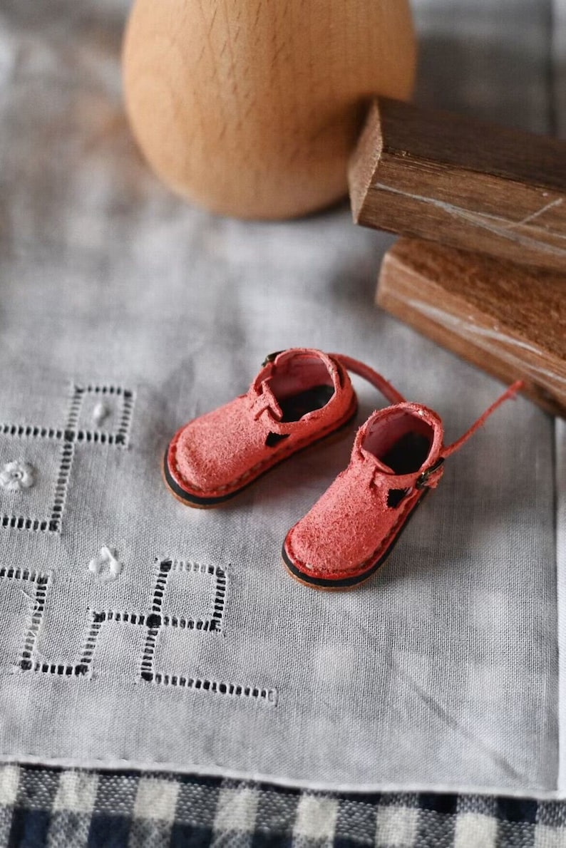 K&ouml;nnte beinhalten: Ein Paar Miniatur-Pinkschuhe aus Leder mit Schnallen. Die Schuhe befinden sich auf einer wei&szlig;en Stoffoberfl&auml;che mit einem zarten Stickmuster.