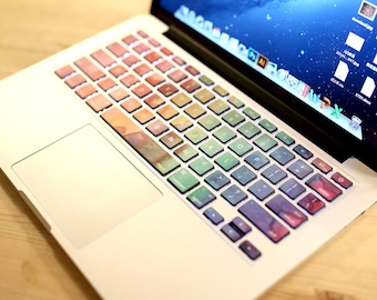 May include: A colorful keyboard sticker set for a Macbook laptop. The stickers feature a watercolor-like design with shades of pink, blue, green, and yellow. The stickers are designed to fit the keys of a Macbook keyboard.