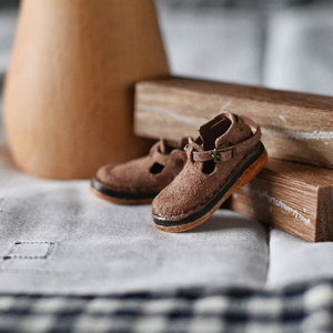 K&ouml;nnte beinhalten: Ein Paar braune Ledersandalen mit einem Schnallenriemen. Die Sandalen liegen auf einem Holzblock und einer wei&szlig;en Stoffoberfl&auml;che.