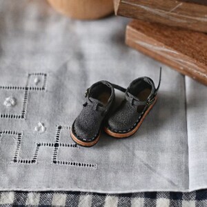 K&ouml;nnte beinhalten: Ein Paar graue Lederschuhe mit braunen Sohlen und einem Schnallenverschluss. Die Schuhe liegen auf einer wei&szlig;en Tischdecke mit einem karierten Muster.