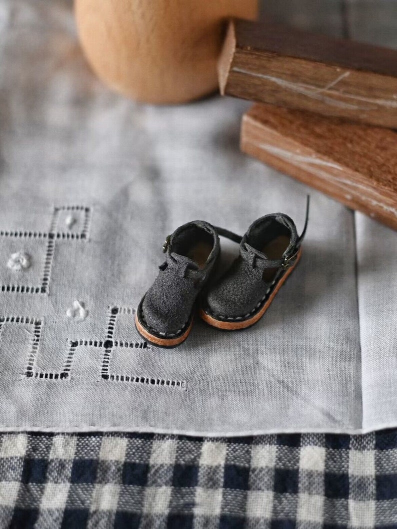 K&ouml;nnte beinhalten: Ein Paar graue Lederschuhe mit braunen Sohlen und einem Schnallenverschluss. Die Schuhe liegen auf einer wei&szlig;en Tischdecke mit einem karierten Muster.