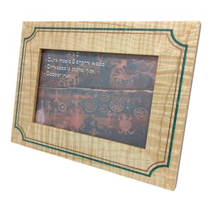 Op de afbeelding: Een houten fotolijst met een lichtgekleurde krullende esdoorn en kersenhouten lijst. De lijst heeft een koperen inleg en een turquoise accent. De lijst bevat een foto met oude tekeningen in petrogliefstijl.