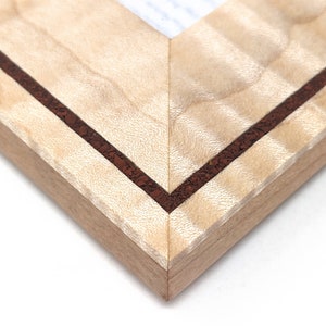 May include: Close-up of a corner of a light wood picture frame with a brown inlay. The frame has a natural wood grain pattern.
