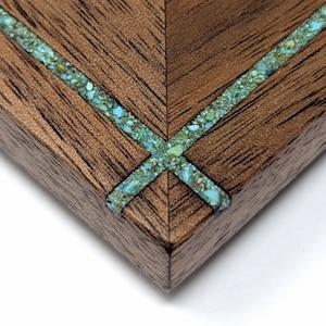 May include: A corner section of a dark brown wooden object featuring a mitered joint. A decorative inlay of crushed turquoise-colored stone runs along the seam of the wood, forming a cross pattern at the corner point of the dark grain surface.