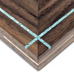 May include: Close-up of a wooden corner with a chevron pattern. The dark brown wood is inlaid with a turquoise-colored stone. The corner has a light-colored border, creating a layered effect. The design is geometric and detailed.