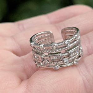 May include: Silver-toned adjustable ring with multiple bands. The ring features a combination of round and rectangular clear stones, and a rope-like texture on some bands. The open design allows for size adjustment.