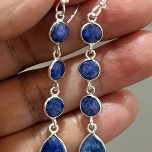 Puede incluir: Un par de pendientes colgantes con cuatro piedras azules engastadas en plata. Cada pendiente consta de tres piedras circulares seguidas de una piedra en forma de lágrima en la base, unidas por eslabones de plata y ganchos de plata.