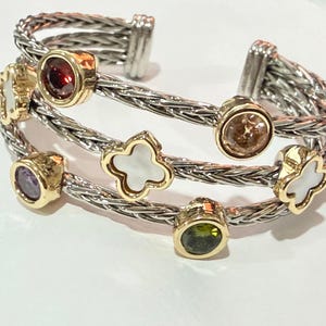 May include: A silver and gold cuff bracelet with three interwoven bands. The bracelet features various gemstones in gold settings, including red, purple, green, and a champagne-colored stone. There are also white clover-shaped accents.