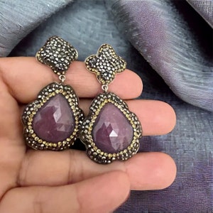 Puede incluir: Pendientes colgantes con una piedra preciosa rosa oscuro facetada en forma de lágrima. Los pendientes tienen una parte superior en forma de flor de color gris oscuro y un borde de pequeñas piedras amarillas. El fondo es una tela drapeada y texturizada.