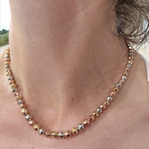 Puede incluir: Collar con cuentas facetadas en tonos oro, plata y oro rosa. El collar se lleva alrededor del cuello, mostrando una mezcla de colores metálicos.