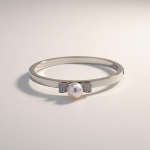 May include: A silver-toned bangle bracelet with a bow-shaped accent. The bow features small, sparkling embellishments and a central, round, light pink pearl. The bracelet is set against a soft, neutral background.