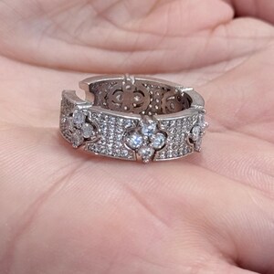 May include: A silver-toned band ring featuring a wide, flat profile with an open back. The surface is densely encrusted with small, clear, sparkling stones and accented with four-petaled floral motifs composed of larger round stones.
