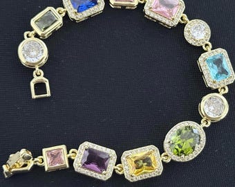 May include: A gold-tone bracelet featuring a series of multi-colored rectangular and oval gemstones, each surrounded by a halo of small clear stones. The gems include blue, pink, light blue, green, purple, and yellow hues.