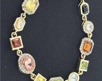May include: Gold-toned bracelet featuring a variety of colorful, faceted gemstones in square, oval, and rectangular shapes. Each gem is set in a gold frame and surrounded by small, clear crystals. The bracelet has a lobster clasp.
