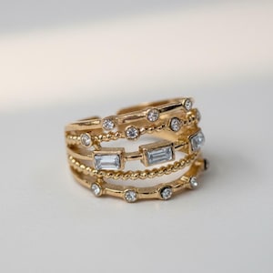 Puede incluir: Un anillo dorado con múltiples bandas, cada una adornada con piedras preciosas claras y brillantes. Algunas bandas presentan pequeñas piedras redondas, mientras que otras tienen piedras de corte rectangular. El anillo tiene un diseño en capas, creando un look audaz y elegante.