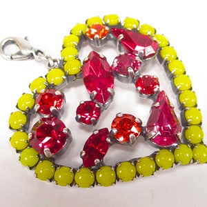 May include: A silver heart-shaped charm with a red and yellow crystal design. The charm has a silver loop for attaching to a bracelet or necklace.