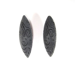 May include: A pair of black, oval-shaped earrings with a carved design. The earrings have a matte finish and are made of a smooth material.